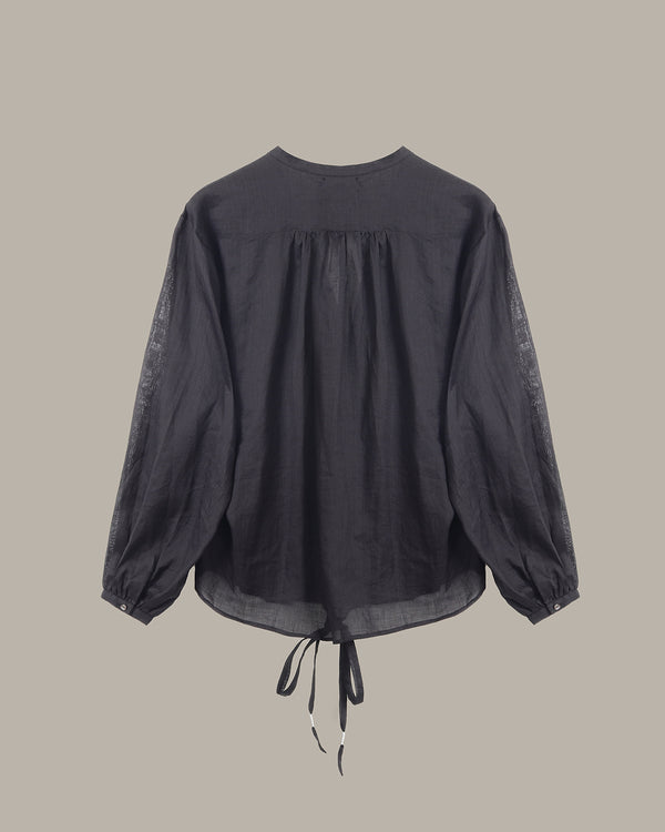 A black long-sleeve blouse with a gathered, pleated back and drawstring ties at the hem, shown against a solid taupe background.