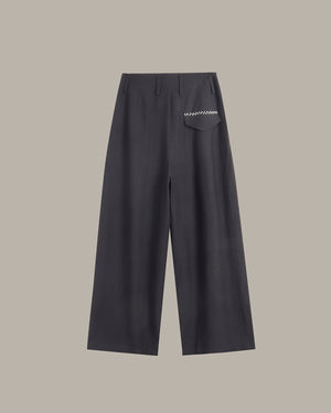 A pair of dark charcoal gray wide-leg trousers with a small white embroidered geometric pattern on the back pocket, shown against a solid taupe background.