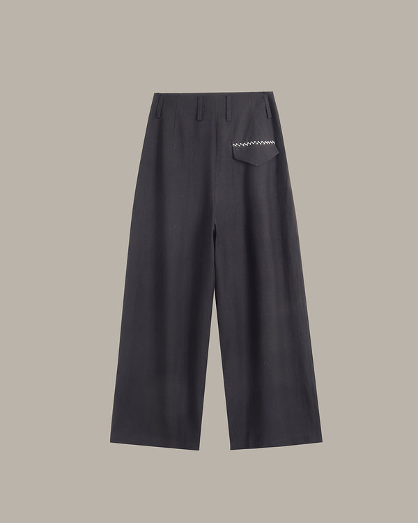 A pair of dark charcoal gray wide-leg trousers with a small white embroidered geometric pattern on the back pocket, shown against a solid taupe background.