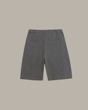 A pair of dark gray tailored shorts shown from the back, featuring a button waistband and two welt back pockets, set against a solid taupe background.