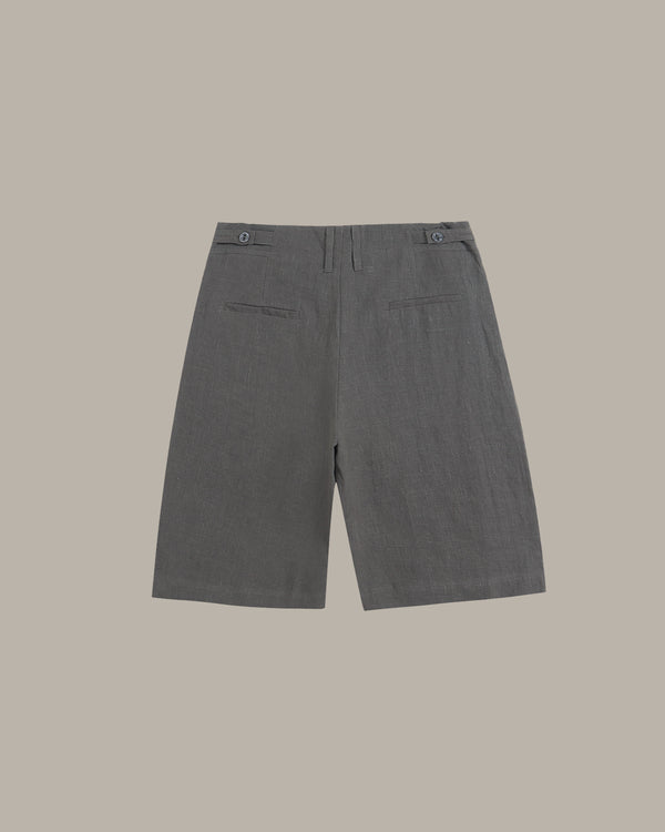 A pair of dark gray tailored shorts shown from the back, featuring a button waistband and two welt back pockets, set against a solid taupe background.
