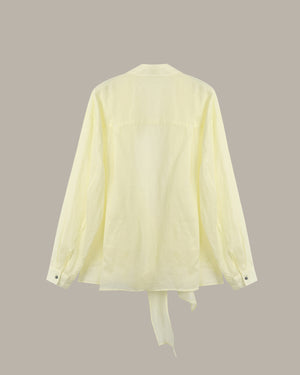 A pale yellow long-sleeve shirt shown from the back, featuring a relaxed fit, buttoned cuffs, and a tie detail at the bottom hem, set against a solid taupe background.
