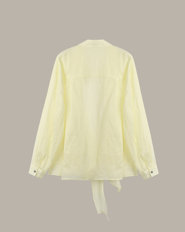 A pale yellow long-sleeve shirt shown from the back, featuring a relaxed fit, buttoned cuffs, and a tie detail at the bottom hem, set against a solid taupe background.