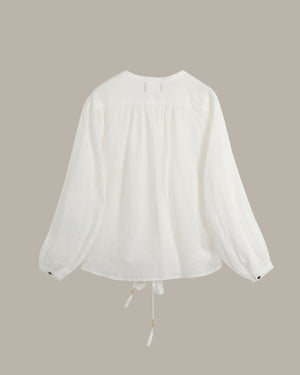 A white long-sleeve blouse with a gathered, smocked bodice and a drawstring tie at the hem, displayed against a solid taupe background.