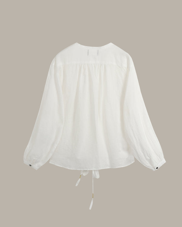 A white long-sleeve blouse with a gathered, smocked bodice and a drawstring tie at the hem, displayed against a solid taupe background.