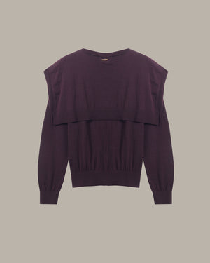 A dark plum purple knit sweater with a layered, asymmetrical ruffle design across the chest and long sleeves, shown flat against a solid taupe background.