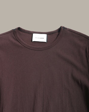 Close-up of a dark brown crewneck t-shirt with a white neck label reading 'COLOR ME' on a solid taupe background.