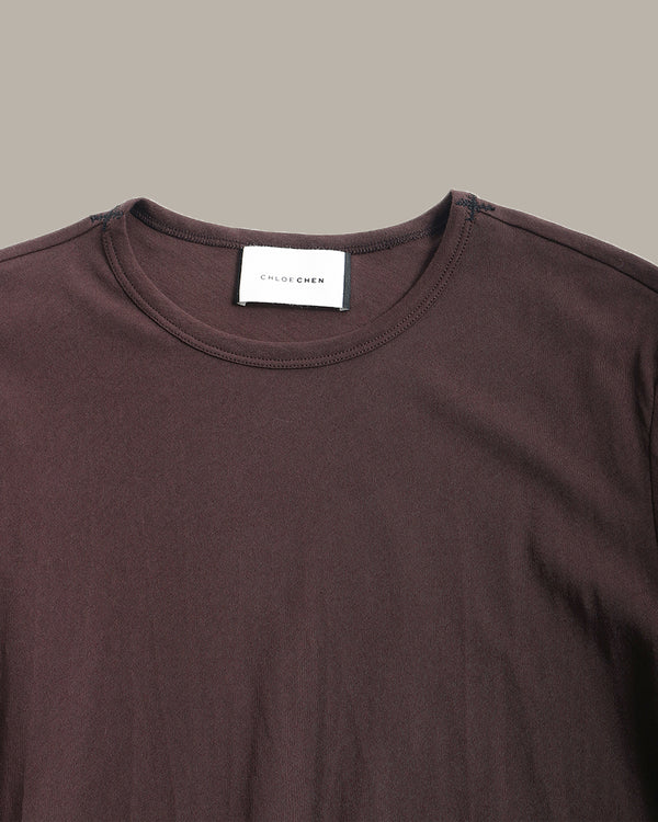 Close-up of a dark brown crewneck t-shirt with a white neck label reading 'COLOR ME' on a solid taupe background.