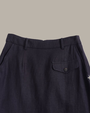Close-up of dark navy trousers showing the waistband, belt loops, front flap pocket with a button, and textured fabric against a solid taupe background.