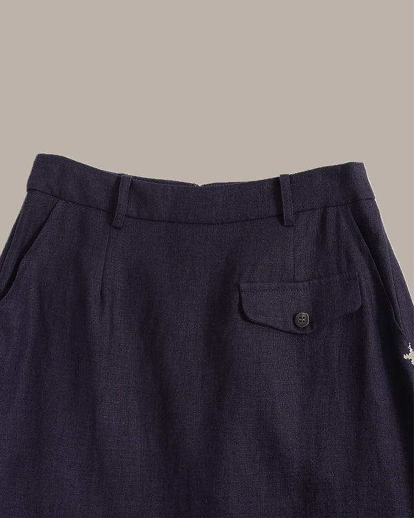 Close-up of dark navy trousers showing the waistband, belt loops, front flap pocket with a button, and textured fabric against a solid taupe background.
