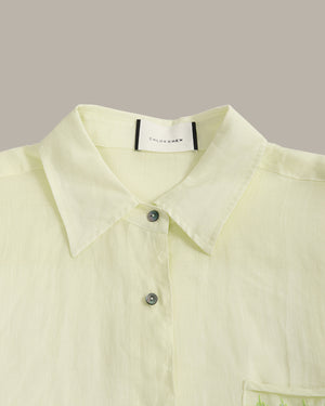 A close-up of a pale yellow-green linen shirt with a pointed collar, silver buttons, and a visible 'COLOR CHEN' brand label inside the collar, set against a solid taupe background.