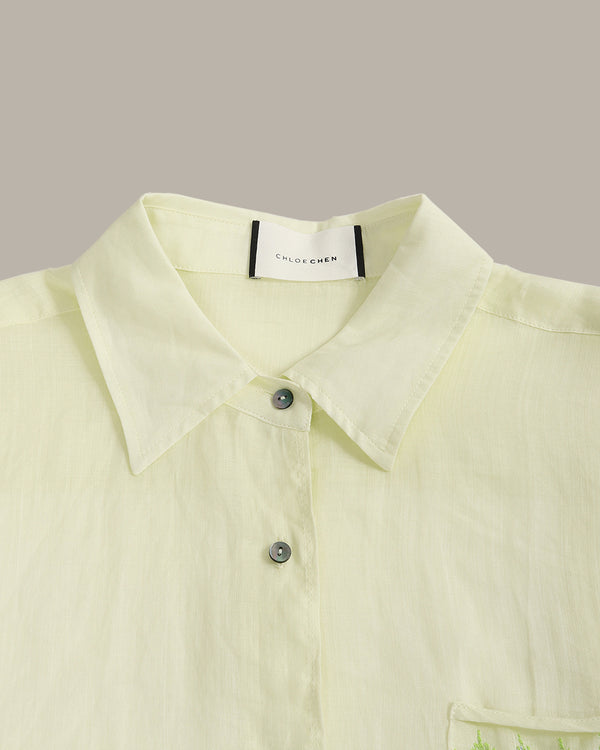 A close-up of a pale yellow-green linen shirt with a pointed collar, silver buttons, and a visible 'COLOR CHEN' brand label inside the collar, set against a solid taupe background.