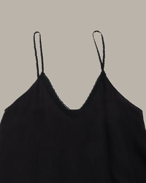 A close-up of a black spaghetti-strap camisole top with a delicate lace trim along the V-neckline, shown against a solid taupe background.