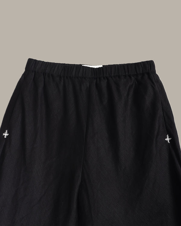 Black wide-leg pants with an elastic waistband, featuring small silver embroidered cross motifs on the left and right thighs, shown against a solid taupe background.