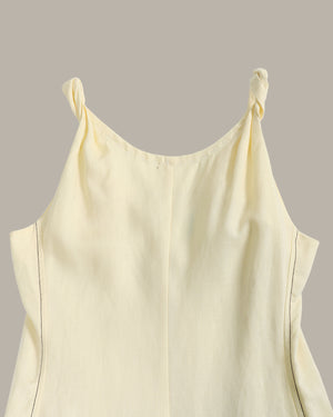 A close-up of a pale cream or off-white sleeveless top with twisted shoulder straps, shown against a solid taupe-gray background.