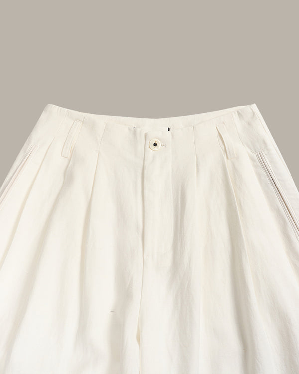 Close-up of a white pleated skirt with a button closure and side pockets, shown against a solid taupe background.