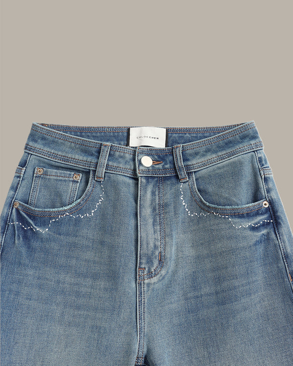 Close-up of a pair of light blue denim jeans showing the front waistband, button closure, belt loops, and decorative white embroidered stitching around the front pockets.