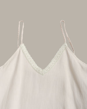 A close-up of a light cream or off-white camisole top with thin spaghetti straps and a delicate floral lace trim along the V-neckline, set against a solid taupe background.
