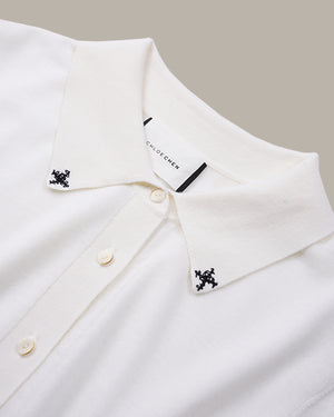 Close-up of a white collared knit top with small black embroidered motifs on both collar points and cream buttons, shown against a light beige background.