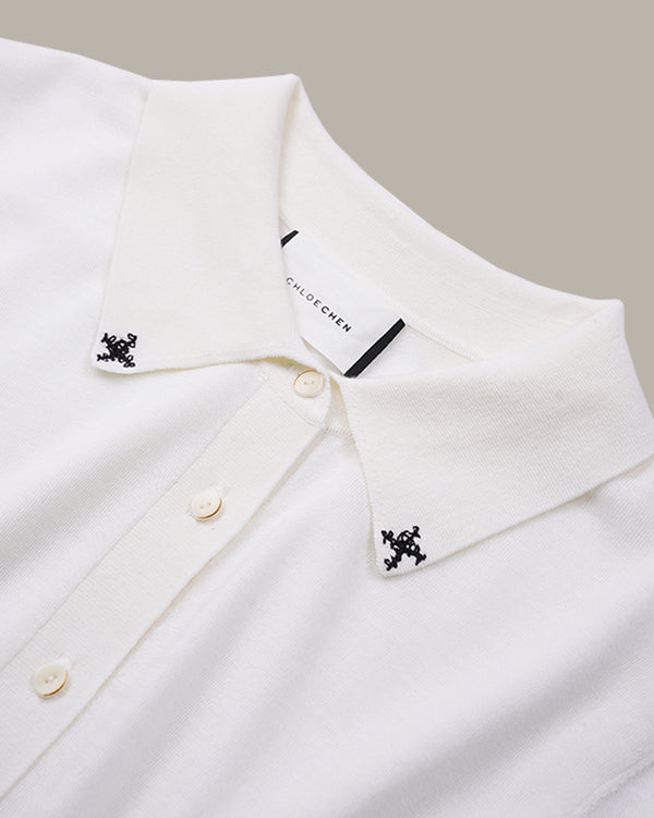 Close-up of a white collared knit top with small black embroidered motifs on both collar points and cream buttons, shown against a light beige background.