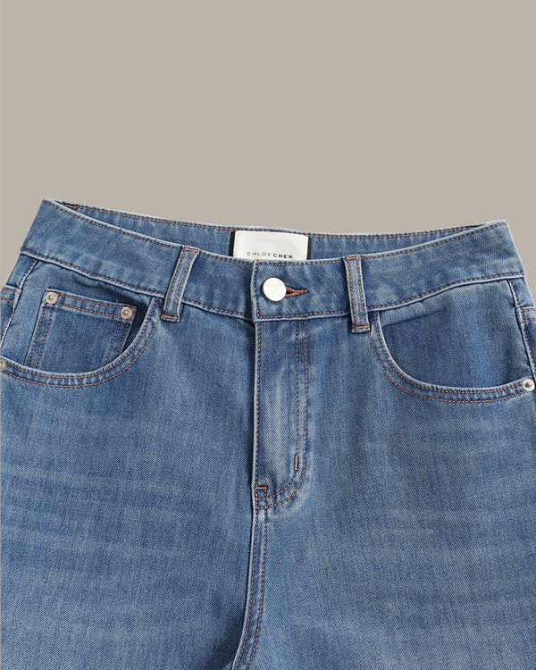 Close-up of the waistband of light blue denim jeans, showing the button closure, belt loops, and front pockets against a solid taupe background.