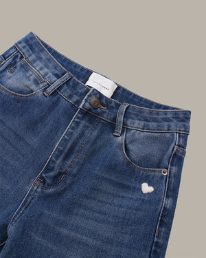 Close-up of a pair of blue denim jeans showing the waistband, belt loops, front pocket, and a small white embroidered heart on the pocket, set against a solid taupe background.