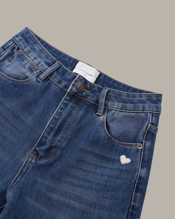 Close-up of a pair of blue denim jeans showing the waistband, belt loops, front pocket, and a small white embroidered heart on the pocket, set against a solid taupe background.