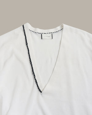 A close-up of a white V-neck top with a black chain trim along the neckline, shown against a solid taupe background.