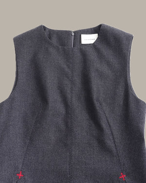 Close-up of the back of a sleeveless dark gray dress with a small white brand label at the neckline and tiny red embroidered details near the bottom corners, shown against a solid taupe background.