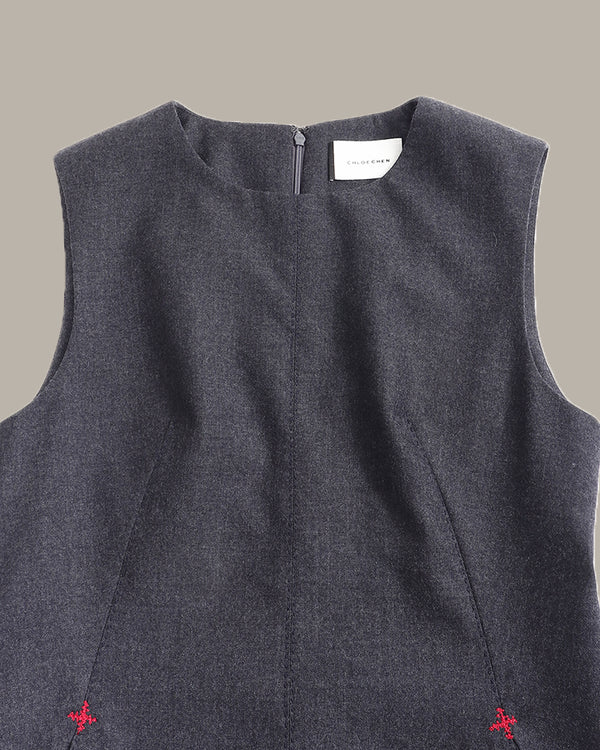 Close-up of the back of a sleeveless dark gray dress with a small white brand label at the neckline and tiny red embroidered details near the bottom corners, shown against a solid taupe background.