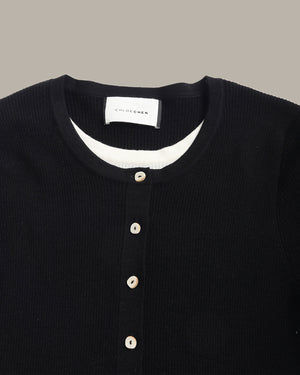 Close-up of a black ribbed knit henley sweater by Chloé, featuring a white inner collar and beige buttons, shown against a solid taupe background.
