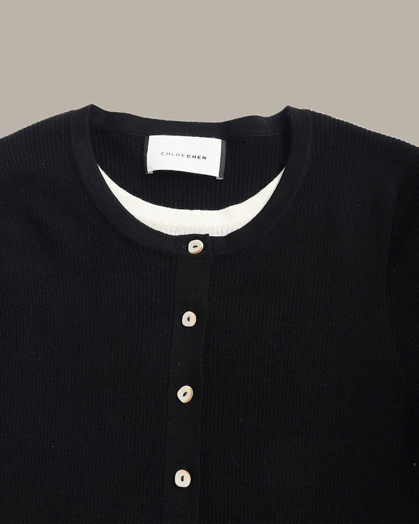 Close-up of a black ribbed knit henley sweater by Chloé, featuring a white inner collar and beige buttons, shown against a solid taupe background.