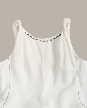 Close-up of a white sleeveless top with thin shoulder straps, black decorative stitching along the neckline, and subtle dark piping on a plain taupe background.
