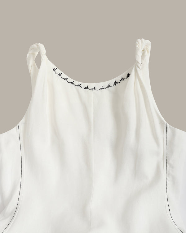 Close-up of a white sleeveless top with thin shoulder straps, black decorative stitching along the neckline, and subtle dark piping on a plain taupe background.