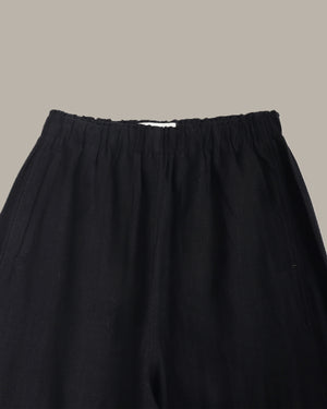 Close-up of black wide-leg trousers showing the gathered elastic waistband and fabric texture against a solid taupe background.