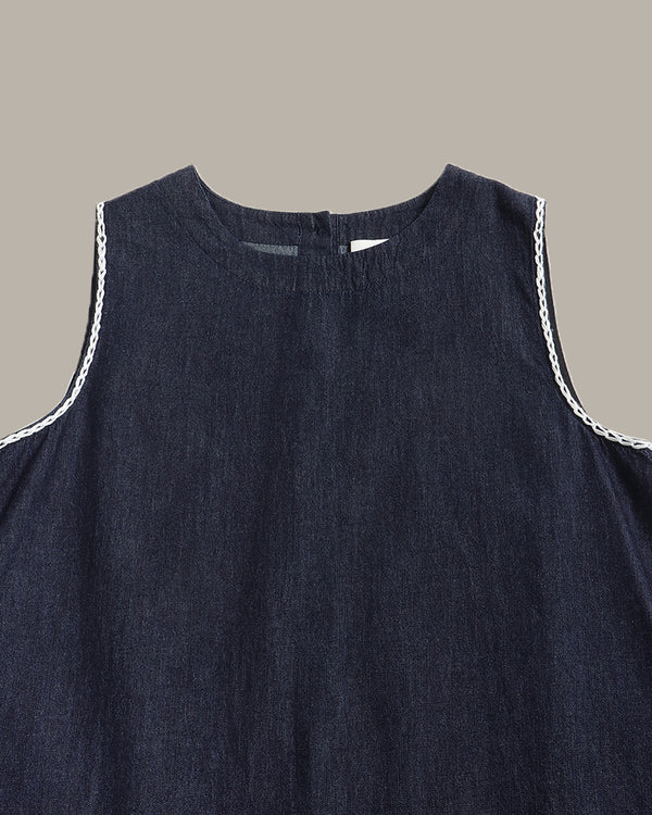 Close-up of a sleeveless dark navy blue top or dress with white braided trim along the armholes, shown against a plain warm gray background.
