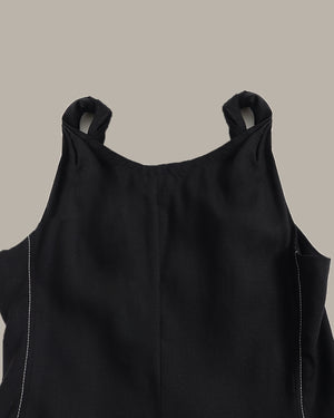 Close-up of a black sleeveless top with shoulder cutouts and white contrast stitching, shown against a solid taupe background.