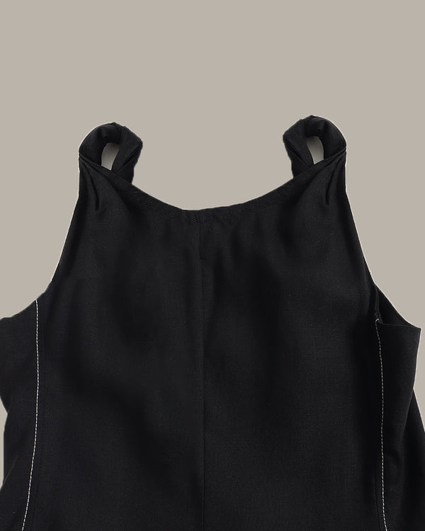 Close-up of a black sleeveless top with shoulder cutouts and white contrast stitching, shown against a solid taupe background.