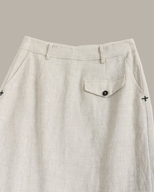 Close-up of a light beige linen skirt showing the waistband, belt loops, a side pocket with a button flap, and small embroidered cross details on the sides.