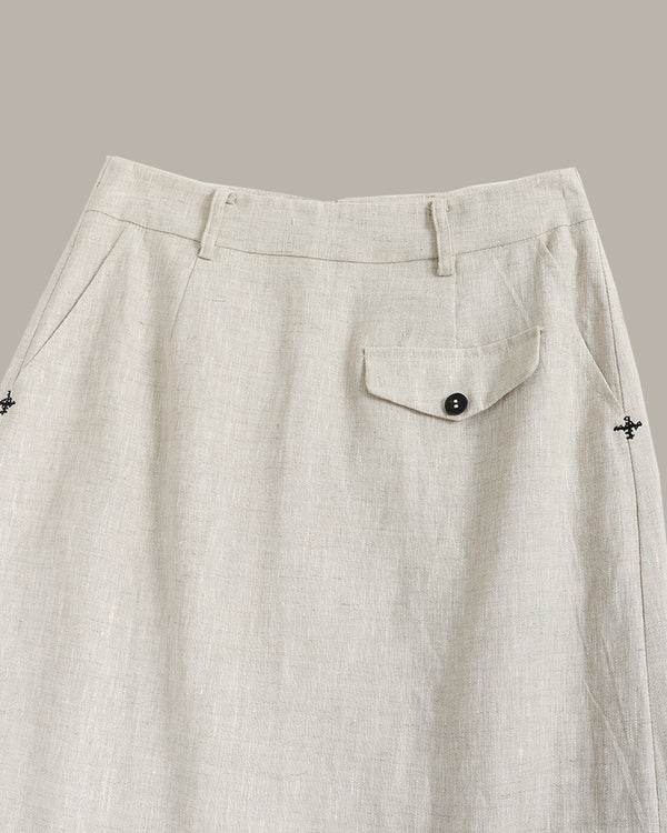 Close-up of a light beige linen skirt showing the waistband, belt loops, a side pocket with a button flap, and small embroidered cross details on the sides.
