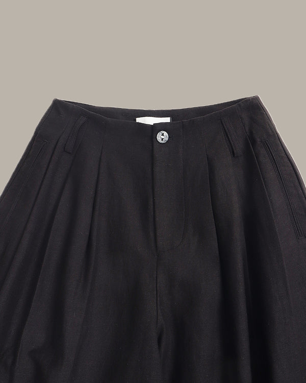 Close-up of a black pleated skirt with a button closure and side slits, shown against a solid taupe background.