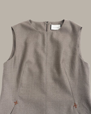 A close-up of a sleeveless taupe-gray linen dress or tunic top with a round neckline, front zipper, and small embroidered floral details near the bottom corners, shown against a solid taupe background.