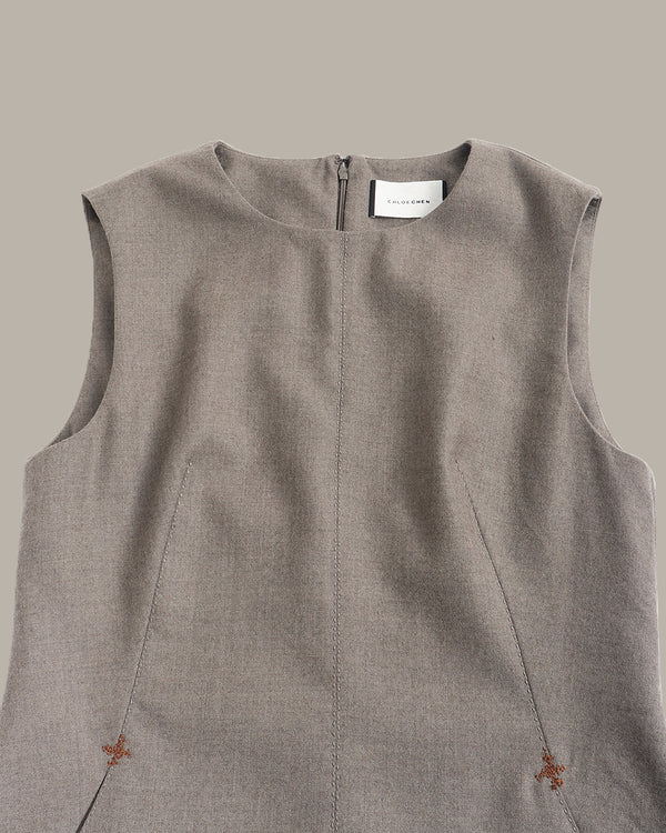 A close-up of a sleeveless taupe-gray linen dress or tunic top with a round neckline, front zipper, and small embroidered floral details near the bottom corners, shown against a solid taupe background.