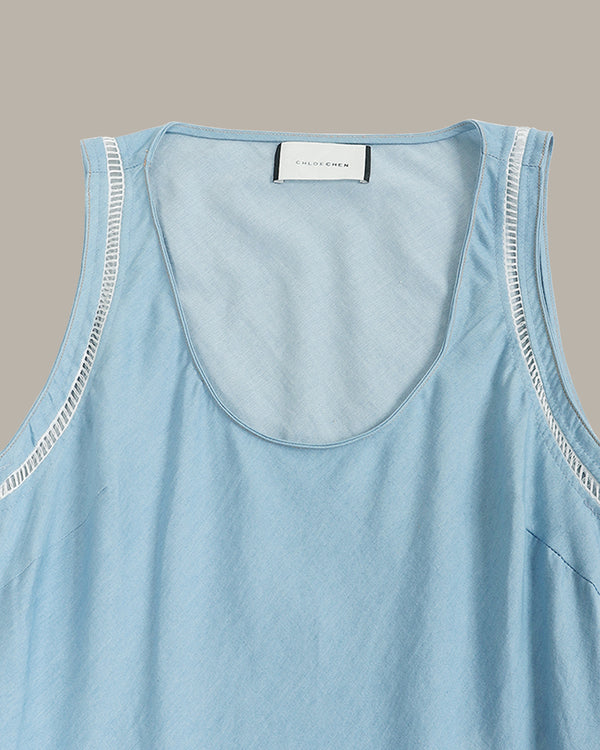 A close-up of a light blue sleeveless top with a wide scoop neckline and decorative white contrast stitching along the armholes, featuring a white brand label at the collar.