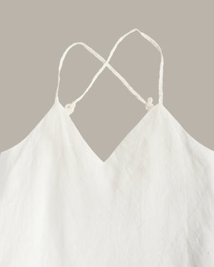 White cotton camisole top with thin adjustable spaghetti straps and knotted back details, shown against a solid taupe background.