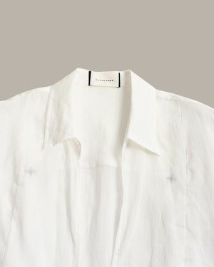 A close-up of a white button-up shirt with a pointed collar, laid flat against a solid taupe background. The brand name 'THE LAUGHERS' is visible on the inner neck label.
