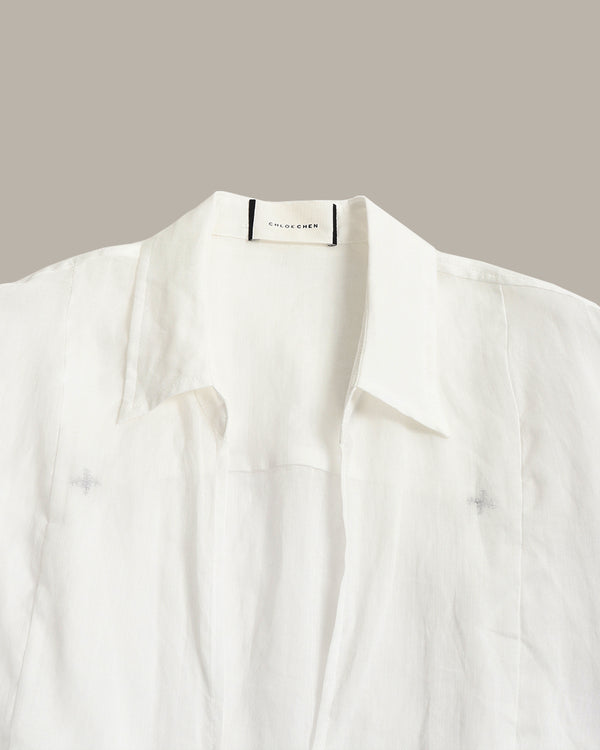 A close-up of a white button-up shirt with a pointed collar, laid flat against a solid taupe background. The brand name 'THE LAUGHERS' is visible on the inner neck label.