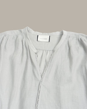 A close-up of a light gray linen blouse with a gathered round neckline, decorative lace trim, and a visible neck label reading 'SILK & STITCH'.