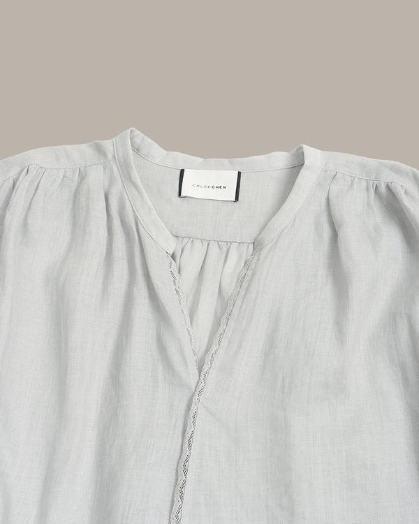 A close-up of a light gray linen blouse with a gathered round neckline, decorative lace trim, and a visible neck label reading 'SILK & STITCH'.