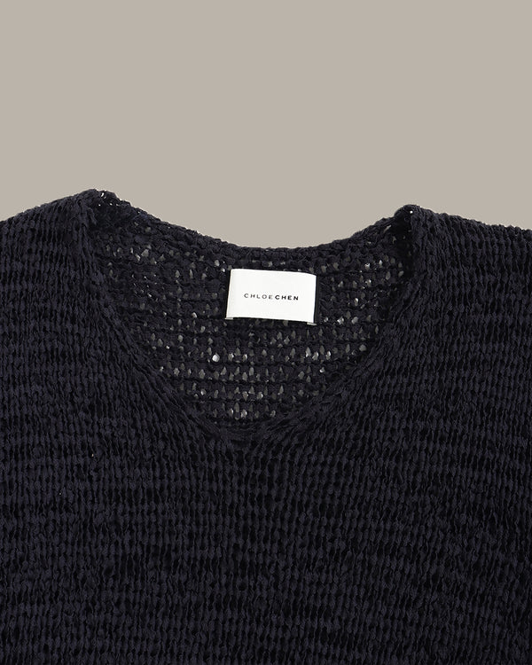 Close-up of a black knit sweater with a V-neck and a visible white neck label reading CHLOE CHEN, set against a plain taupe background.